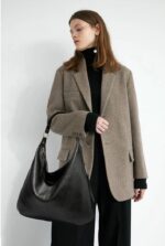 Delaney Large Hobo Bag-black (2) Woman wearing a gray blazer and black turtleneck while carrying a large black Delaney Hobo Shoulder Bag with a relaxed slouchy design