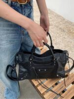 Black Ridley Rivet Motorcycle Bag carried by the top handles, featuring a structured shape, front zip pocket, rivets and long shoulder strap