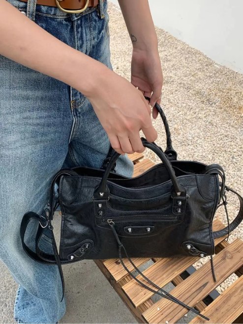 Black Ridley Rivet Motorcycle Bag carried by the top handles, featuring a structured shape, front zip pocket, rivets and long shoulder strap