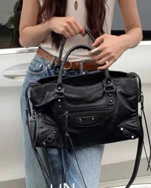 Large black Ridley Rivet Motorcycle Bag held by the top handles, showing front zipper pocket, buckle accents, rivets and shoulder strap