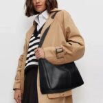 Sutton Hobo Shoulder Bag-black (3) Woman wearing black Sutton Hobo Shoulder Bag with adjustable strap and slouchy design, styled with a beige trench coat and striped sweater