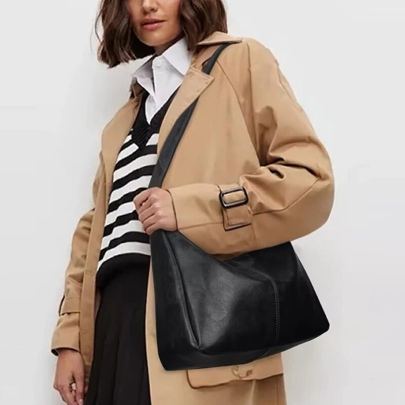 Woman wearing black Sutton Hobo Shoulder Bag with adjustable strap and slouchy design, styled with a beige trench coat and striped sweater