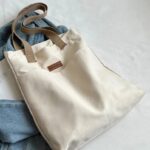Tegan Large Canvas Tote Bag-white (2) White Tegan Large Canvas Tote Bag laid flat on blue denim jeans, showing its soft canvas texture, beige straps and front pocket design