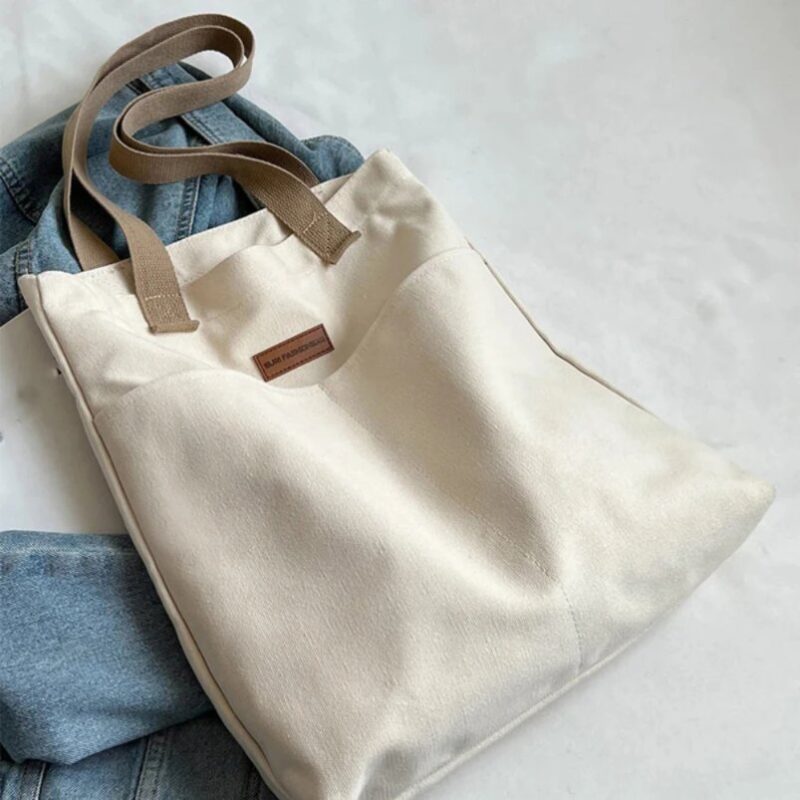 White Tegan Large Canvas Tote Bag laid flat on blue denim jeans, showing its soft canvas texture, beige straps and front pocket design