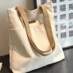 Tegan Large Canvas Tote Bag-white (6) White Tegan Large Canvas Tote Bag standing on a black table with long beige shoulder straps, two front pockets and brown label